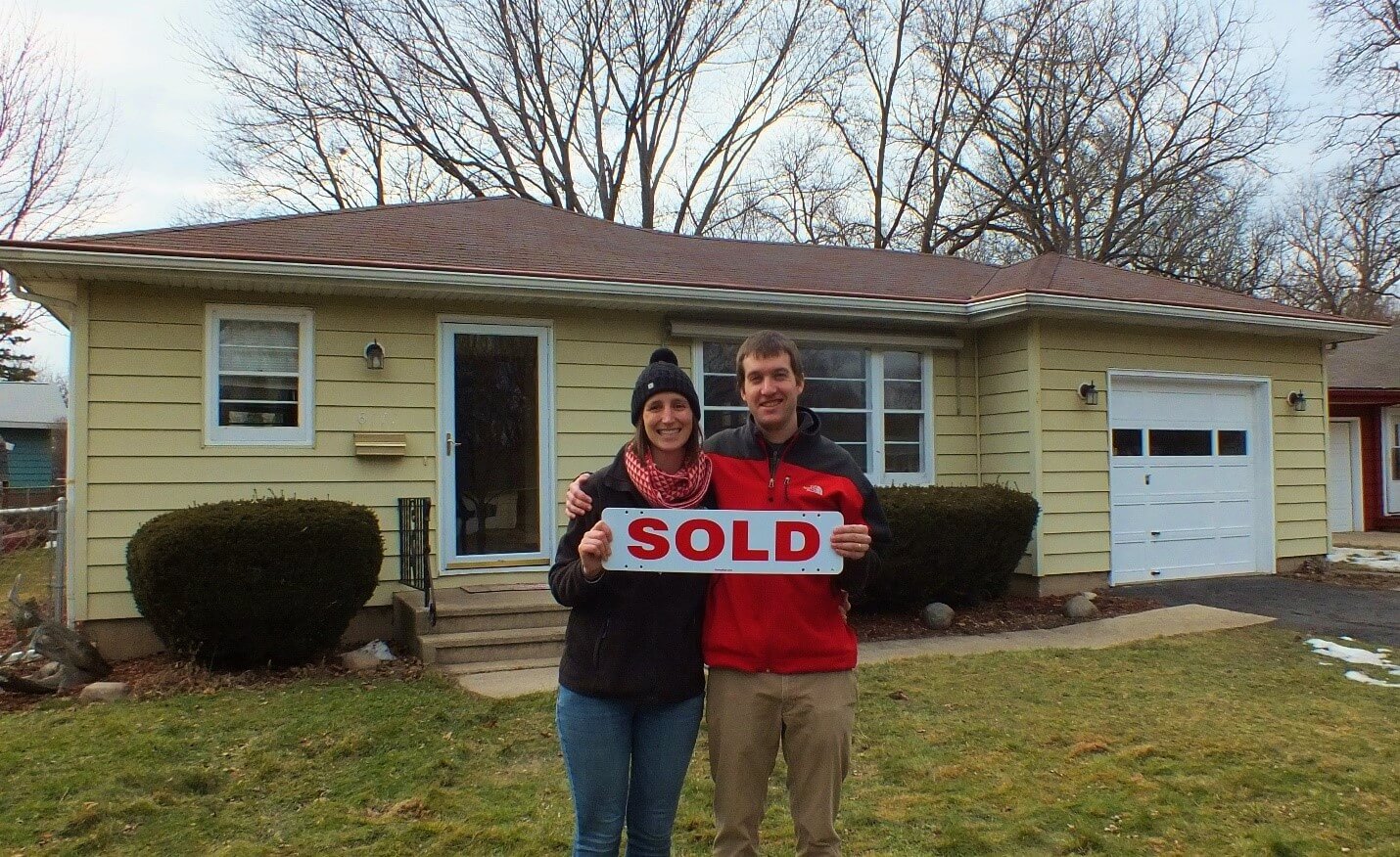 man and woman standing out side of yellow house with sold sign